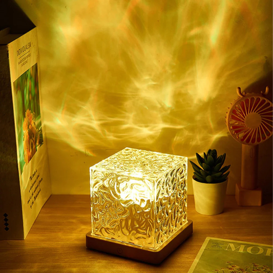 The Tren-Dora CrystalWave™ Ripple Lamp sits on a wooden desk, casting swirling golden patterns. Nearby are a potted succulent, a small circular fan, an open magazine, and a standing card with a floral image.