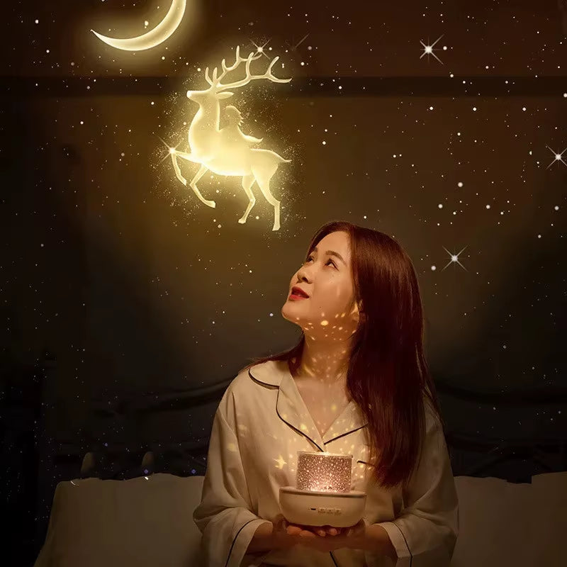 A woman in pajamas sits on her bed, smiling up at golden reindeer and a crescent moon projected by the Tren-Dora StarGlow™ Rotating Galaxy Projector Lamp, which fills the dark wall with dreamy stars for a magical atmosphere.